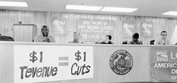 Panelists at town hall meeting, a banner states 1 dollar in revenues for each dollar in cuts