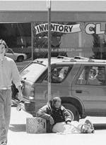 Ronie sleeping on the street leaning next to a pole, as young man walks by.